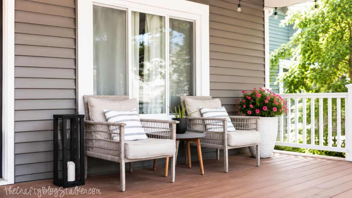Front porch with two wicker chairs, striped pillows, a small side table, black lantern, and pink flowers beside a gray house.