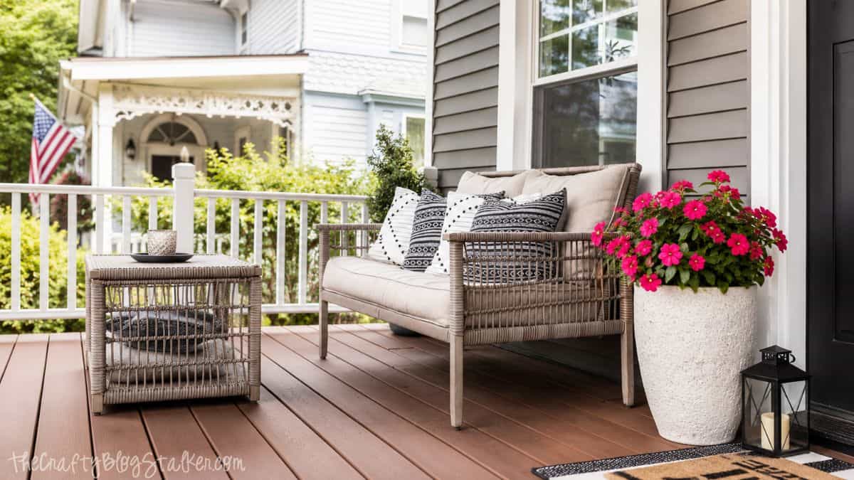 Cozy front porch with wicker seating, patterned pillows, pink flower planters, and a black lantern beside a gray house.