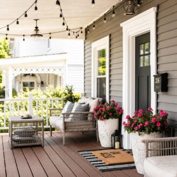 Covered front porch with wicker seating, string lights, pink flower planters, lanterns, and a striped welcome mat outside a gray house.
