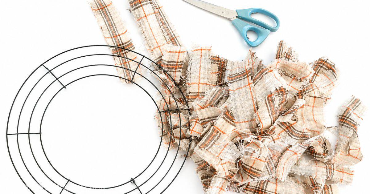 Supplies for a DIY fabric wreath, including a wire wreath form, plaid fabric strips, blue-handled scissors, and tied fabric knots on a white surface.