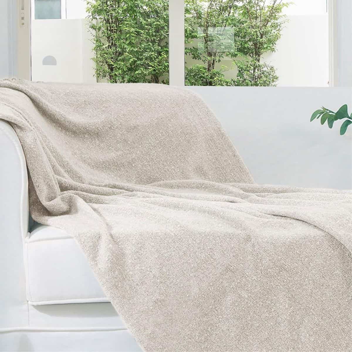 Soft cream knit throw blanket draped over a white sofa in a bright room with large windows and green plants.