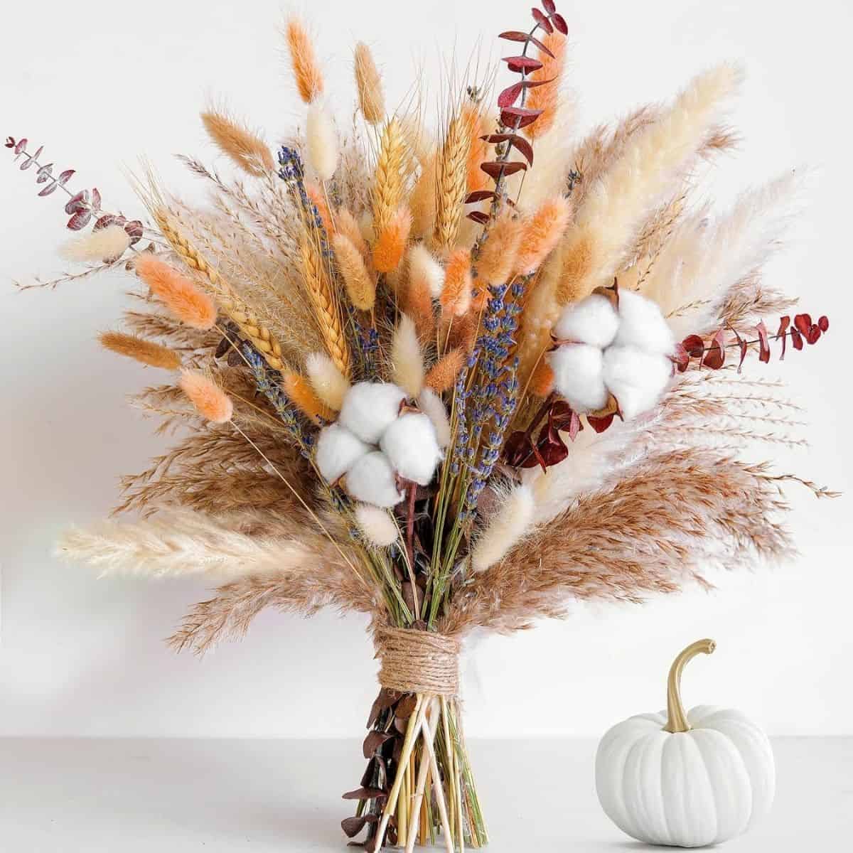Fall bouquet of dried wheat, grasses, lavender, eucalyptus, and cotton stems tied with twine beside a small white pumpkin.