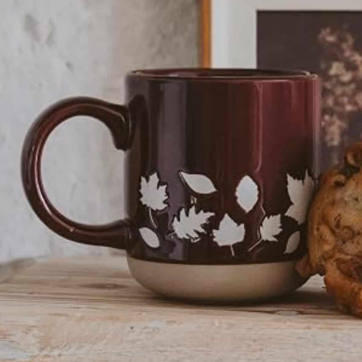Burgundy ceramic coffee mug with cream fall leaf designs and a beige base, displayed on a rustic wooden surface.