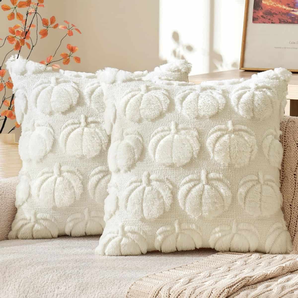Two cream textured throw pillows with raised pumpkin patterns displayed on a neutral sofa with a knit blanket.