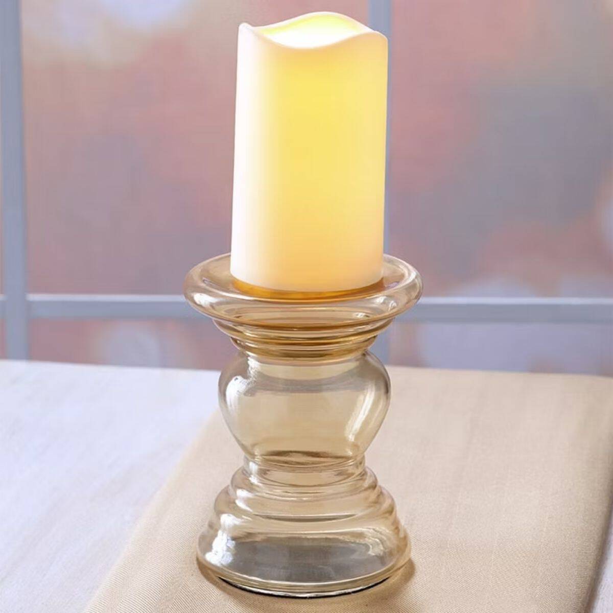 Glowing ivory pillar candle displayed on a tall clear glass candleholder on a table near a window.