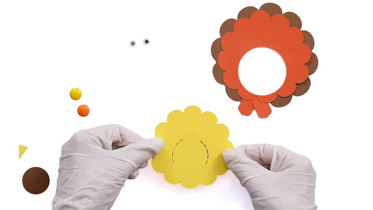 Hands holding the yellow cardstock layer with a perforated opening while assembling the turkey candy treat holder.