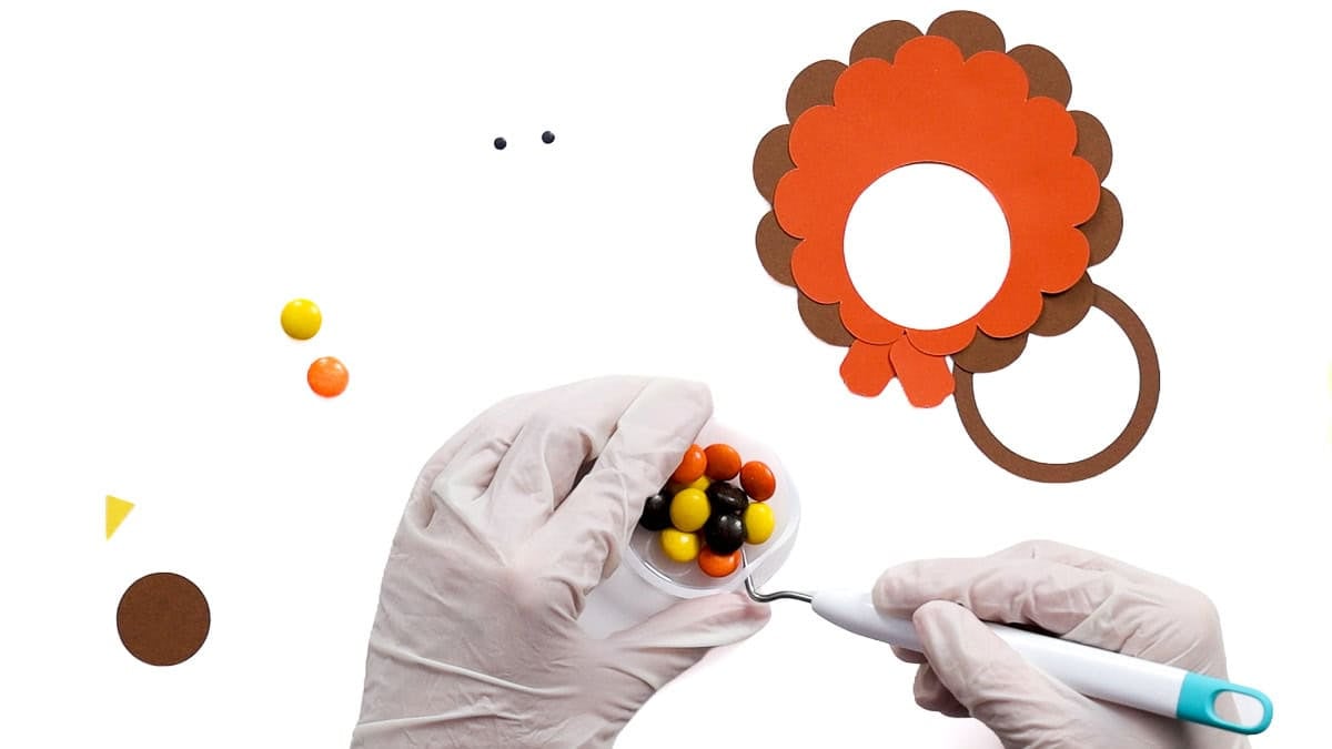 Hands removing the adhesive backing from a clear candy dome filled with Reese's Pieces for a cardstock turkey treat holder.