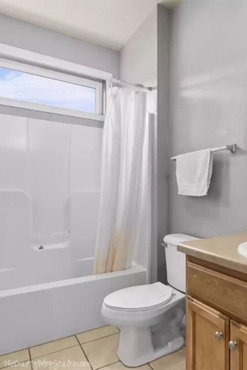 Small bathroom with beige tile floor, white bathtub and toilet, light wood vanity, gray walls, and a white shower curtain stained at the bottom.