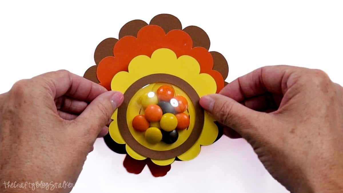 Hands attaching a brown cardstock ring around the candy-filled dome on a layered paper turkey treat holder.