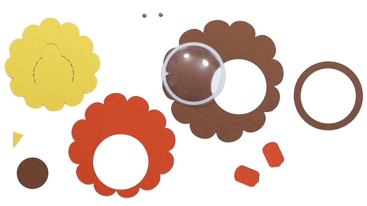Cut cardstock pieces, googly eyes, and a clear plastic dome laid out for assembling a layered turkey candy treat holder.