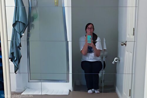 A girl sitting on a toilet across from a mirror wall.