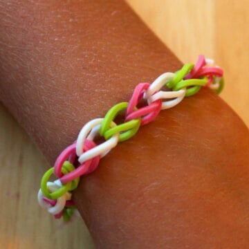 A close-up of a colorful rubber band bracelet in pink, white, and green, woven in a chain design and worn on a wrist.