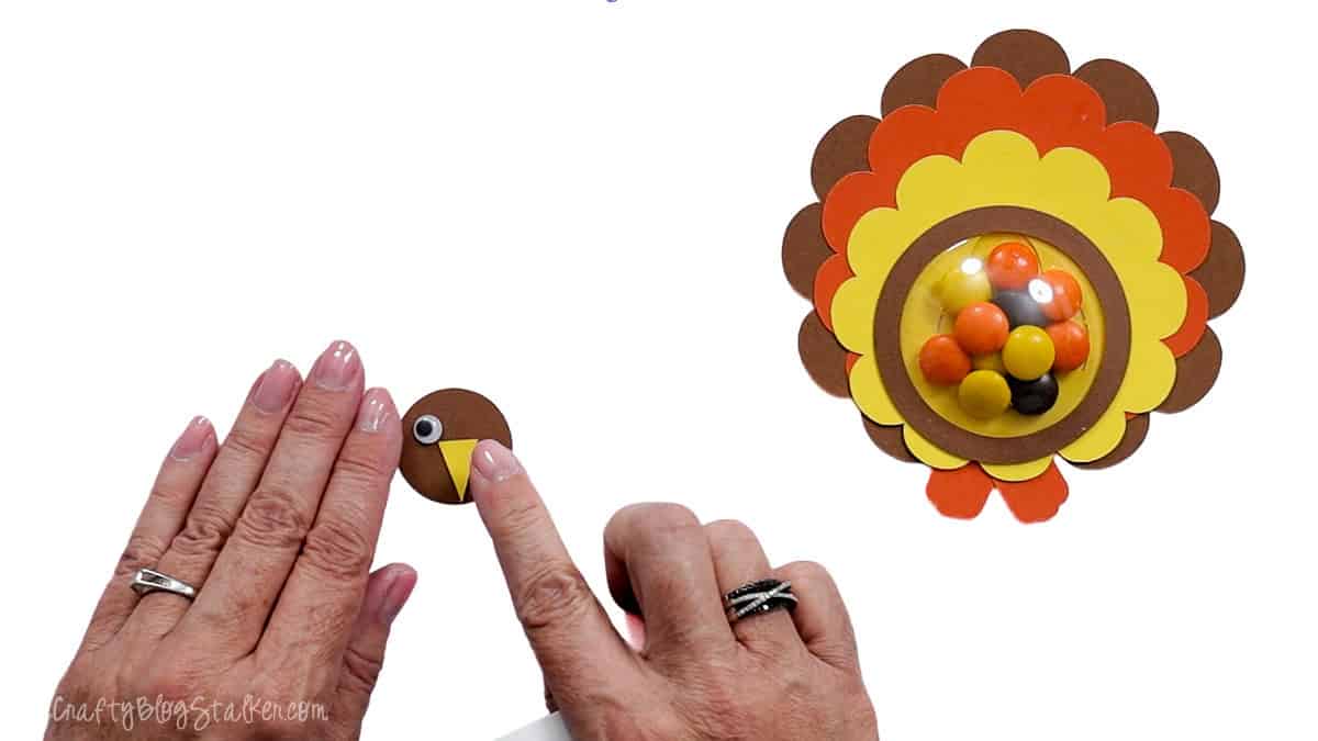 Hands assembling the paper turkey face with googly eyes and a yellow beak beside the finished candy-filled turkey treat holder.