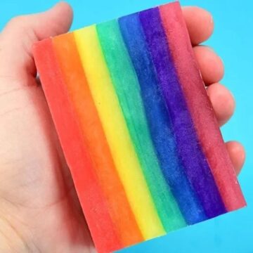 Hand holding a colorful rainbow-striped bar of soap with vibrant layers of red, orange, yellow, green, blue, and purple against a bright blue background.