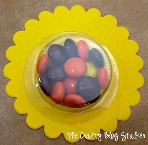 Reeces Pieces in a plastic treat cup, adhered to a paper yellow scallop circle.