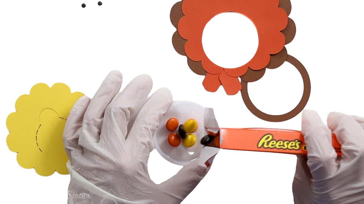 Hands pouring Reese's Pieces into a clear plastic dome while assembling a layered cardstock turkey candy treat holder.