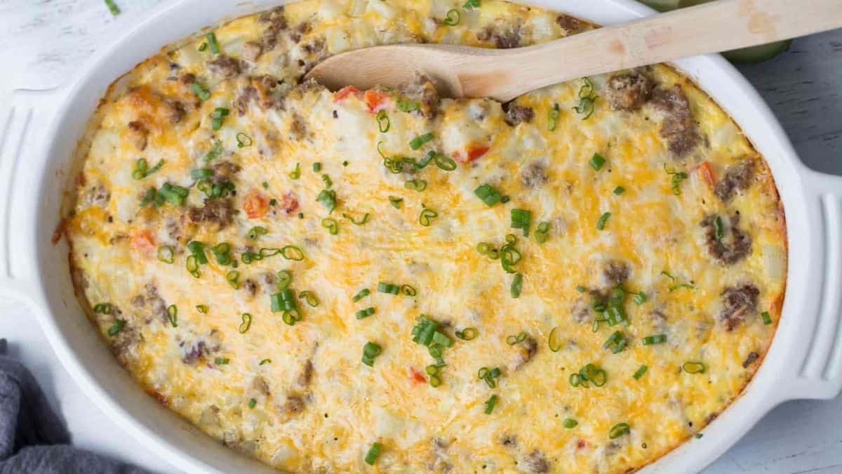 Baked sausage and potato breakfast casserole topped with melted cheese and chopped green onions in a white dish.