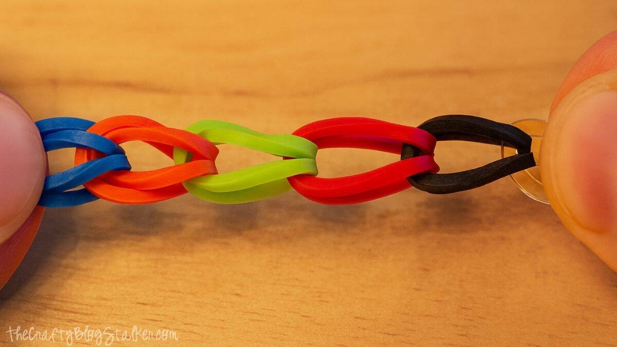 Close-up of a simple loom band bracelet in progress, featuring red, green, orange, blue, and black rubber bands linked together between two fingers.