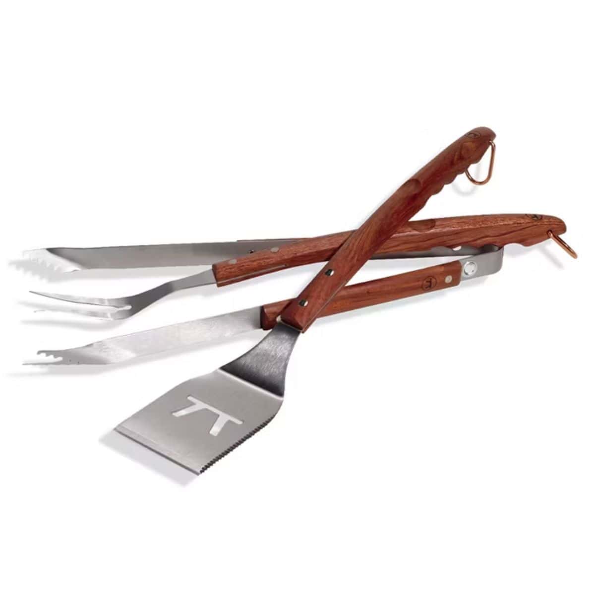 Set of stainless steel BBQ tools with wooden handles, including tongs, a fork, and a spatula with a cutout design, displayed on a white background.