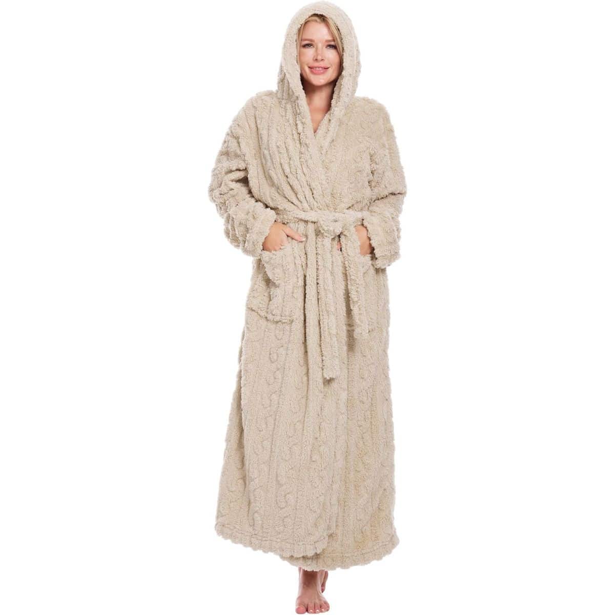 Woman wearing a long beige hooded bathrobe with a belt and front pockets, standing barefoot and smiling.