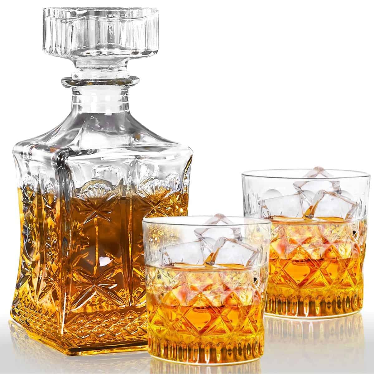 Crystal whiskey decanter filled with amber liquor and two matching glasses with ice cubes, displayed on a reflective surface against a white background.