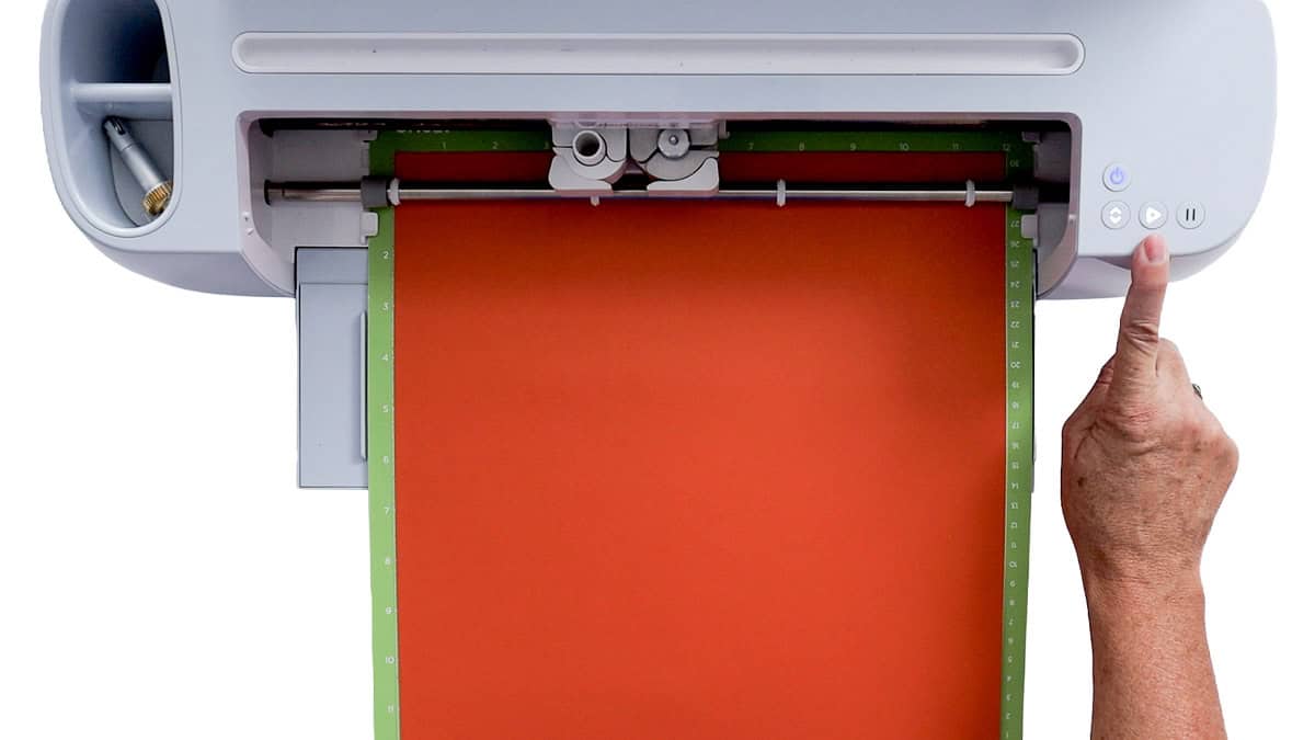 Cricut cutting machine with orange cardstock loaded on a green cutting mat as a hand presses the start button.
