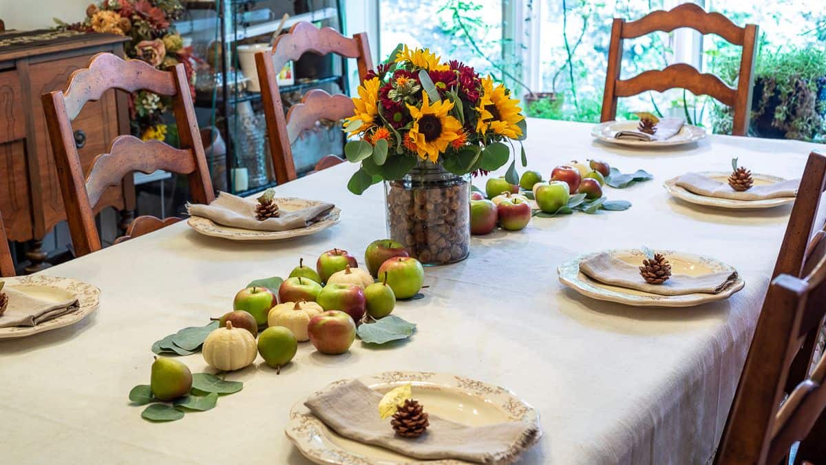 Fall table with a sunflower and walnut centerpiece, surrounded by apples, mini pumpkins, and pinecone napkin holders on cream-colored plates.