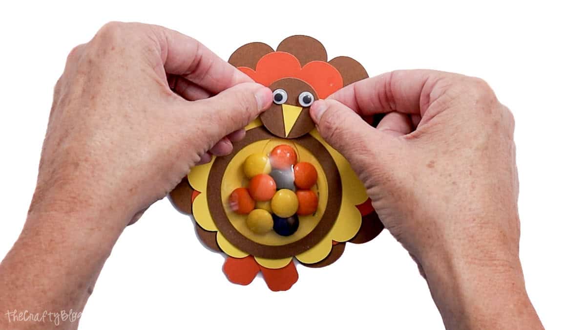 Hands attaching the paper turkey face to a colorful candy-filled turkey treat holder made with layered cardstock.