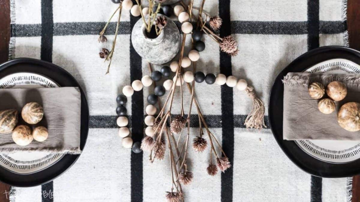 Modern fall tablescape with a black and white plaid table runner, wooden bead garland, dried florals, and small gourds on neutral napkins and plates.