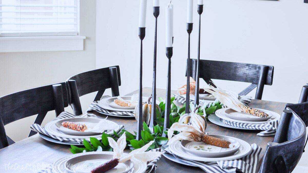 Modern fall tablescape with striped napkins, white plates, Indian corn accents, tall black candlesticks, and green leaf garland centerpiece.