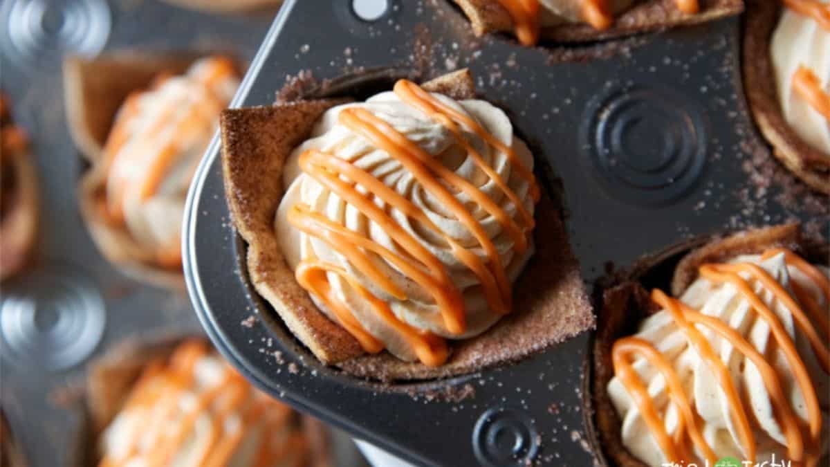 Pumpkin mousse tortilla cups topped with whipped cream and drizzled with orange icing, shown in a muffin tin ready to serve.