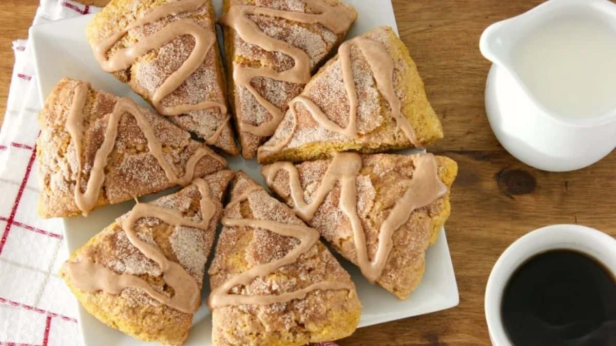 Plate of pumpkin snickerdoodle scones drizzled with cinnamon glaze, served with a cup of coffee and a small pitcher of cream on a wooden table.