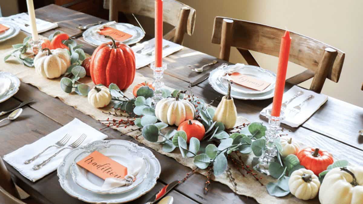 Fall tablescape with white and orange pumpkins, greenery, and red taper candles arranged on a wooden table set with white plates and linen napkins.