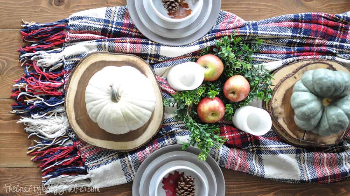 Cozy fall tablescape with pumpkins, apples, greenery, and candles arranged on a plaid blanket runner with wood slice chargers and neutral dishes.