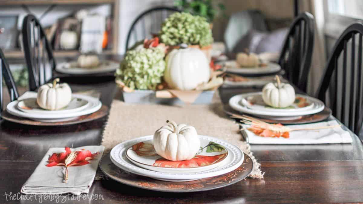 Fall farmhouse table setting with white plates, mini pumpkins, burlap runner, and a centerpiece of white pumpkins and hydrangeas for Thanksgiving decor.