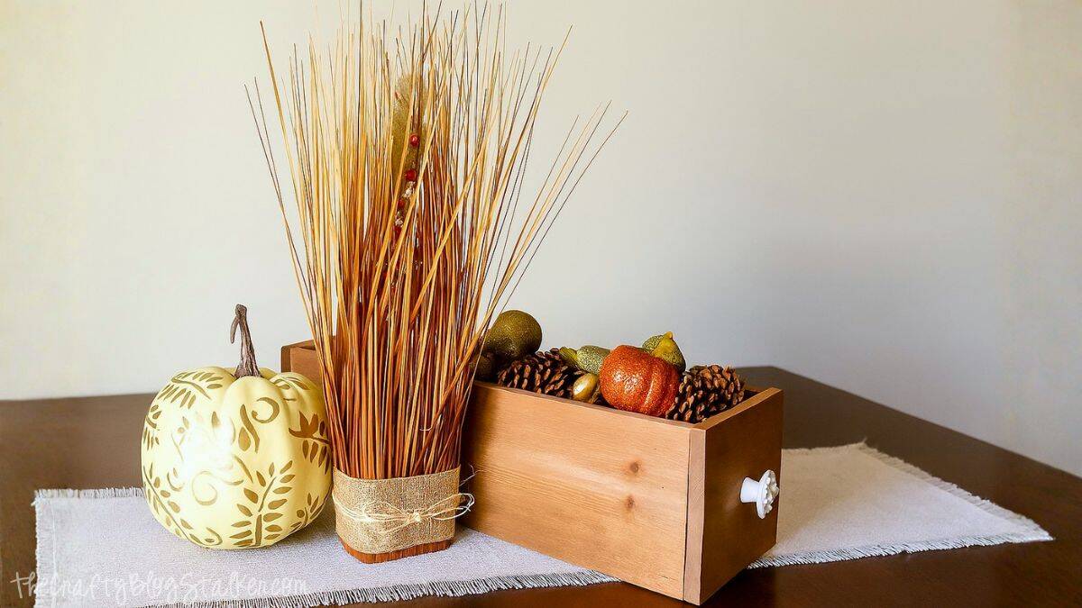 DIY Thanksgiving centerpiece with a wooden box filled with pumpkins, pinecones, and fall decor, paired with a gold pumpkin and tall wheat arrangement on a table.