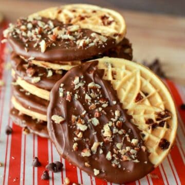 Stack of chocolate chip pizzelle cookies dipped in chocolate and topped with chopped nuts, placed on a red and white striped surface with scattered chocolate chips.
