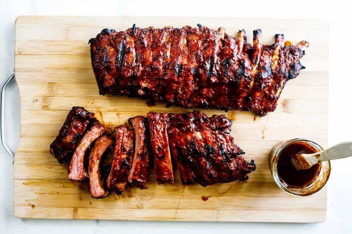 Juicy grilled baby back ribs coated in BBQ sauce, sliced and served on a wooden cutting board with a jar of extra sauce and basting brush on the side.