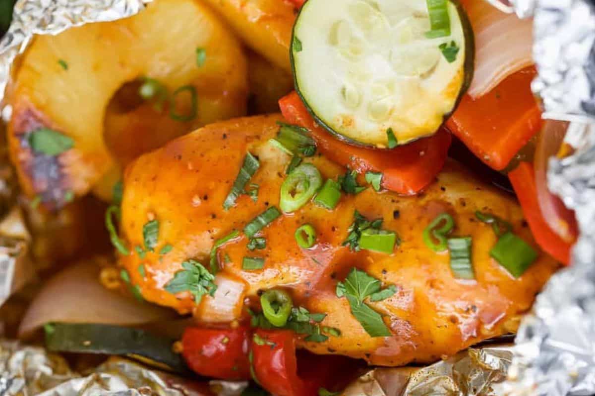 Grilled BBQ Hawaiian chicken cooked in foil with zucchini, bell peppers, onion, and pineapple slices, topped with chopped green onions and herbs.