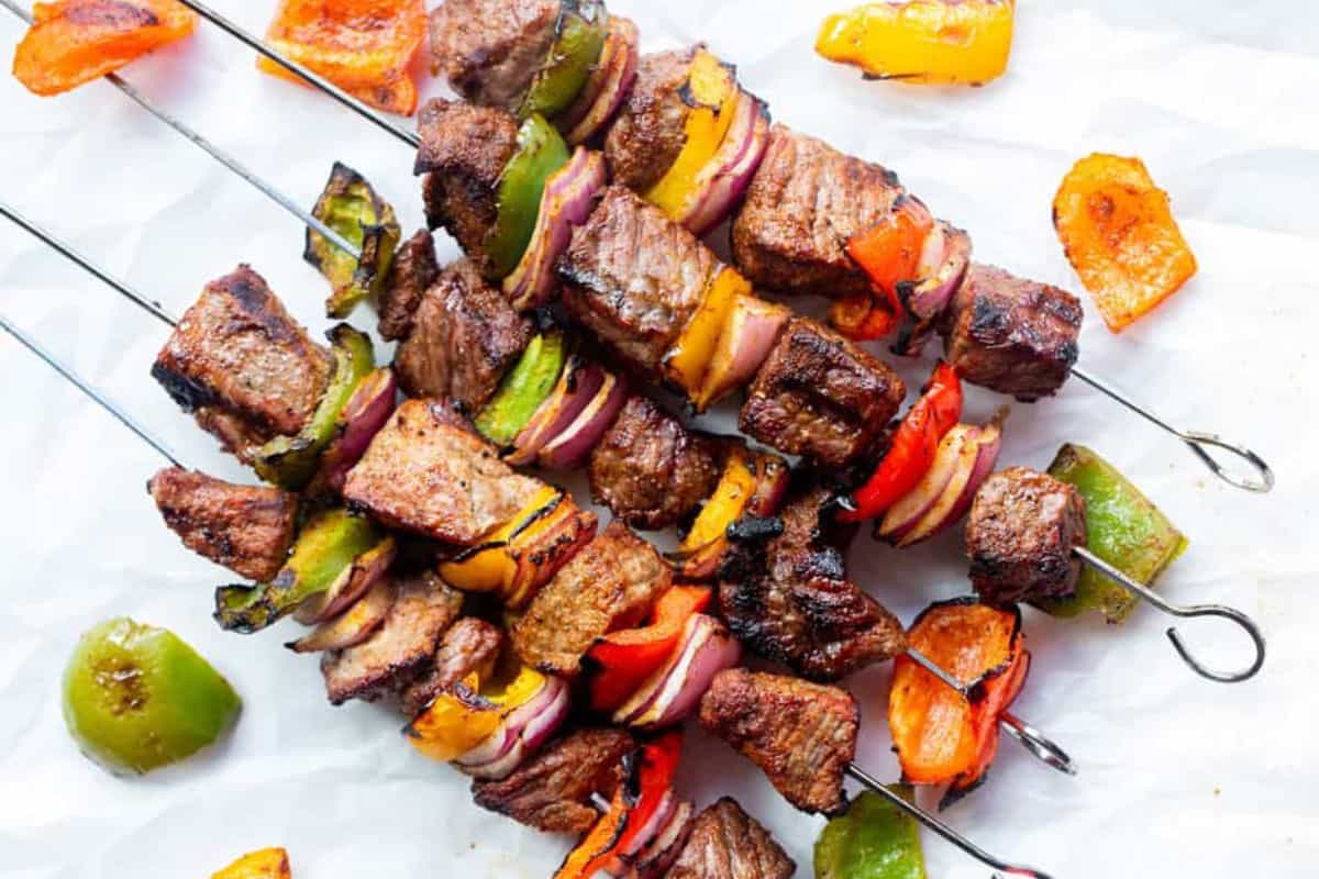 Grilled steak fajita kebabs with juicy beef, bell peppers, and red onions on metal skewers, charred and ready to serve on parchment paper.