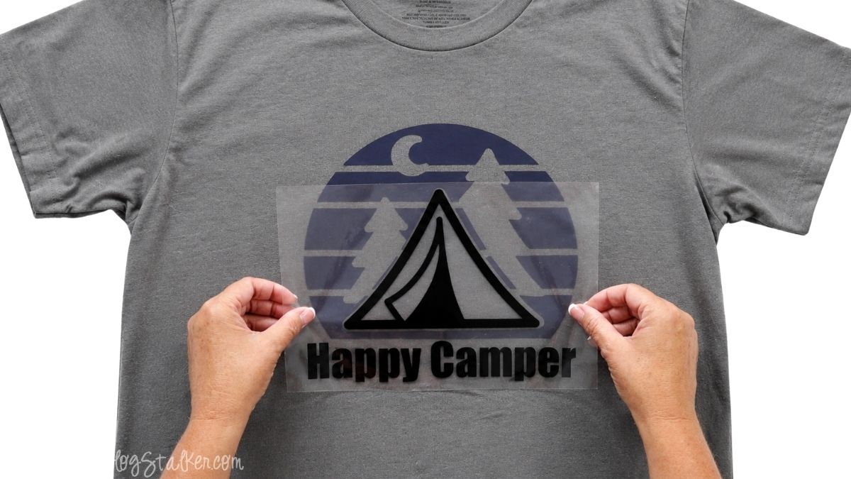Hands positioning the black tent and Happy Camper iron-on vinyl layer over the navy background on a gray T-shirt.