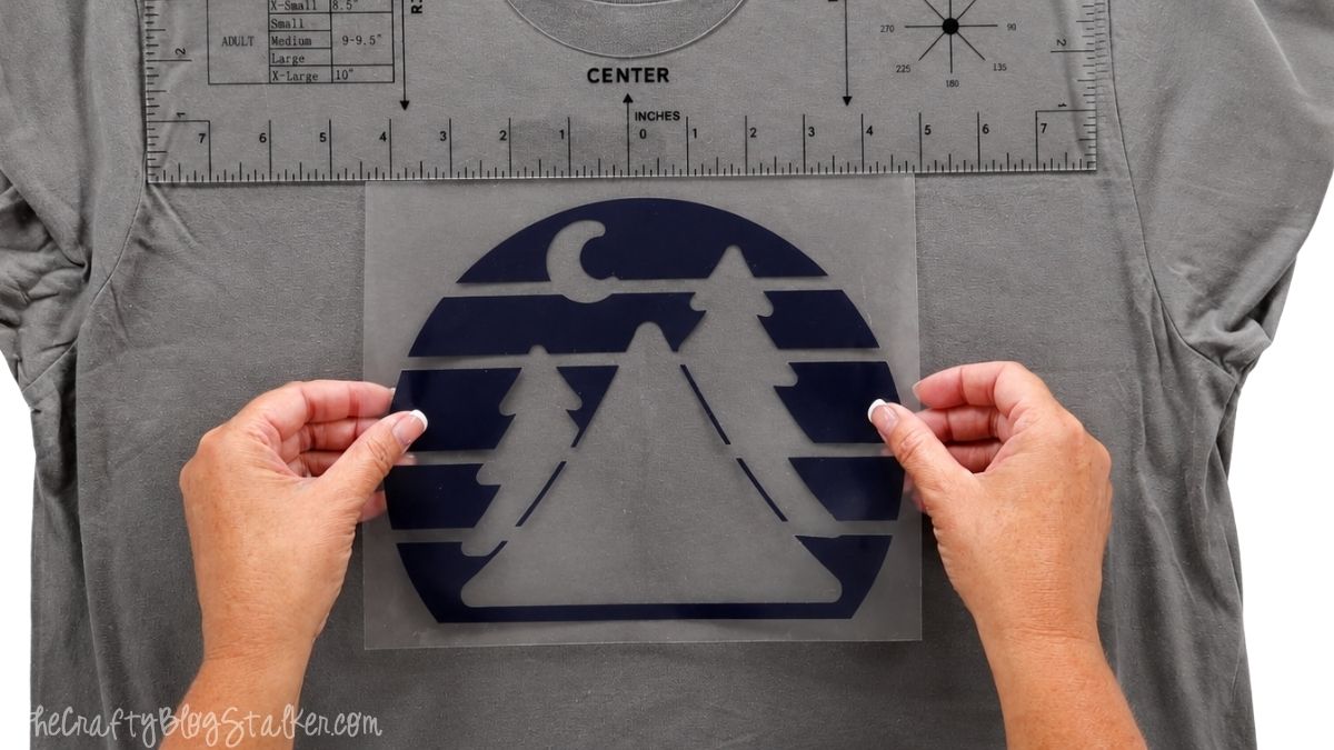 Hands positioning the navy iron-on vinyl layer of a Happy Camper design on a gray T-shirt using a shirt alignment ruler.