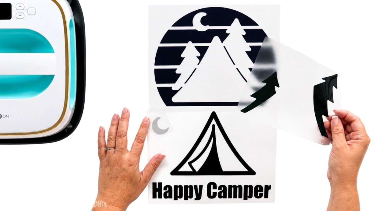 Hands separating layers of a Happy Camper iron-on vinyl design with a tent, pine trees, moon, and striped background beside a heat press.