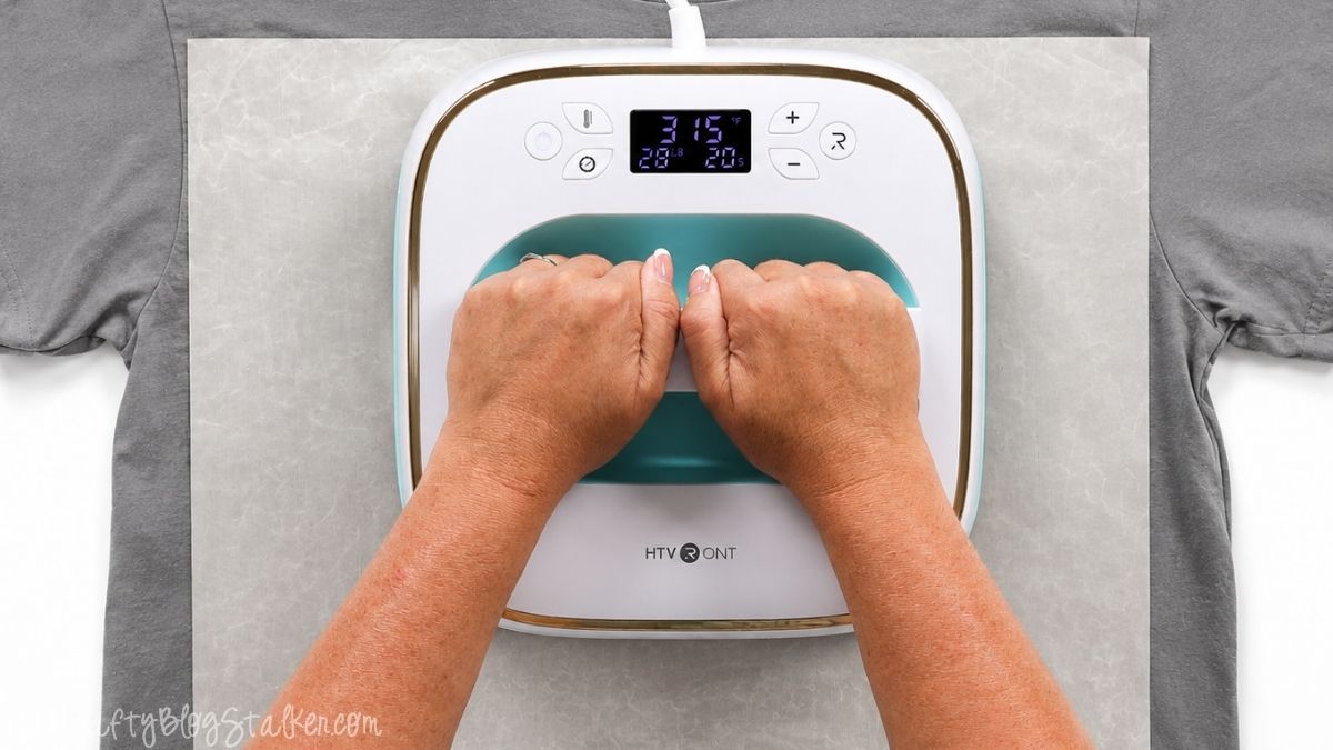 Hands pressing an HTVRONT heat press onto a gray T-shirt to apply layered iron-on vinyl, with protective paper covering the design.