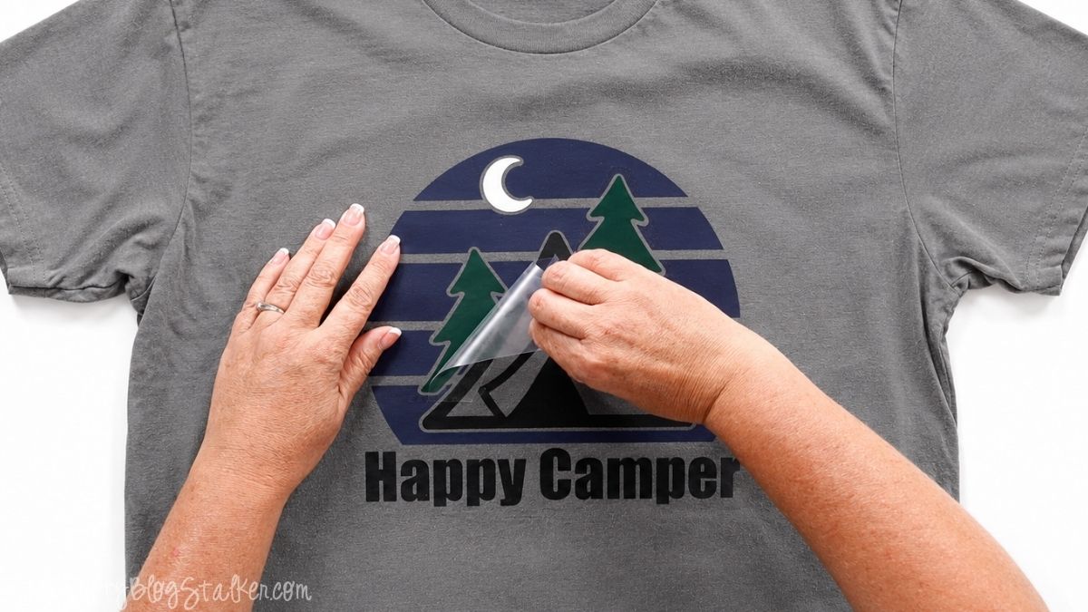 Hands peeling the carrier sheet from a layered Happy Camper iron-on vinyl design applied to a gray T-shirt.