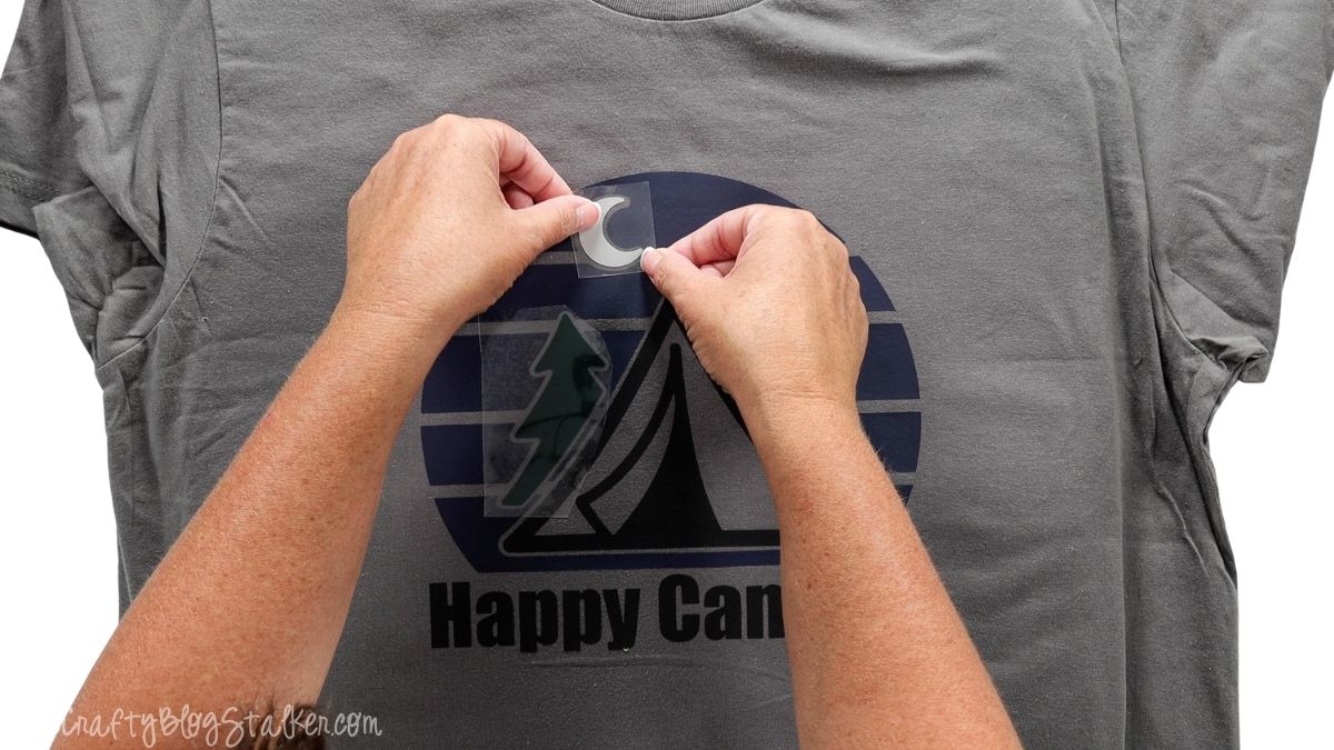 Hands positioning the white crescent moon iron-on vinyl layer on a gray Happy Camper T-shirt with a tent and pine tree design.