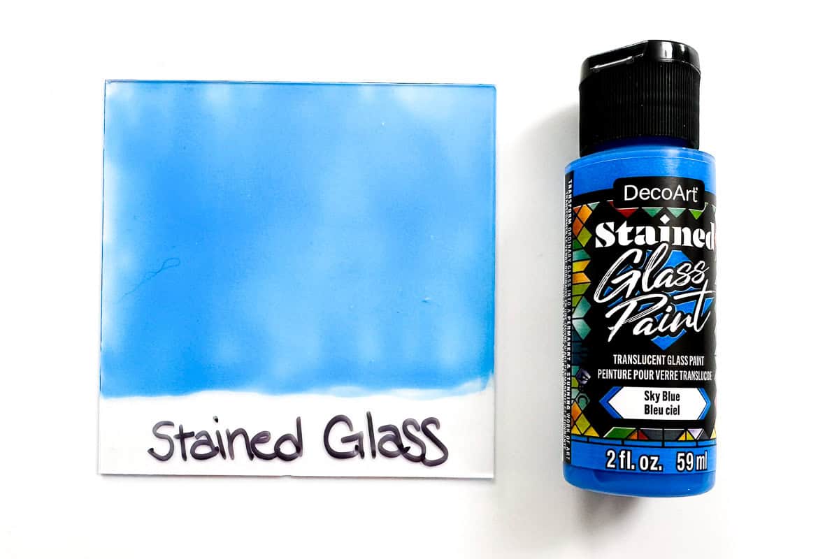 A bottle of Deco Art blue stained glass paint next to a glass pane painted with the same paint.