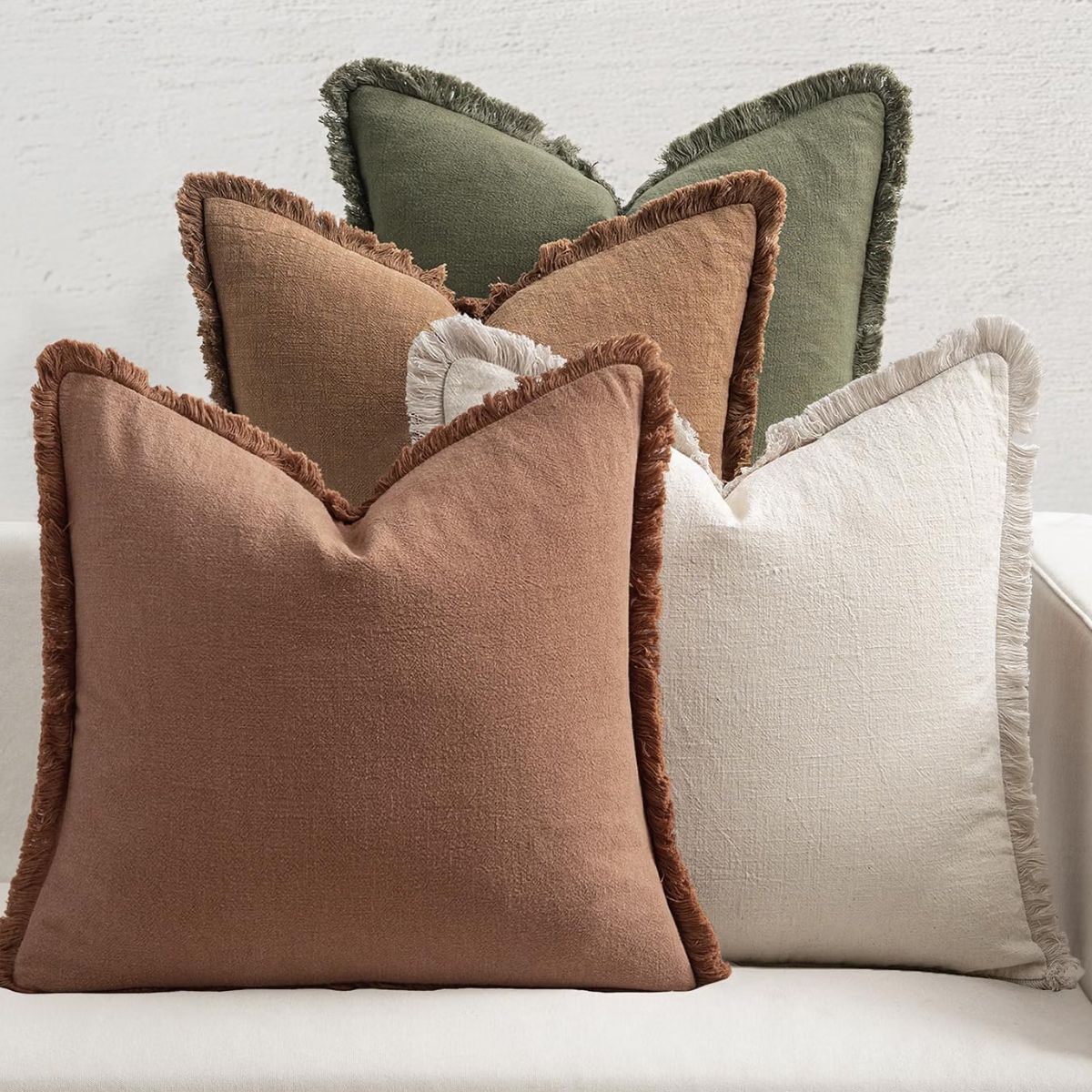 Neutral fringe throw pillows in rust brown, cream, and olive green arranged on a white sofa.