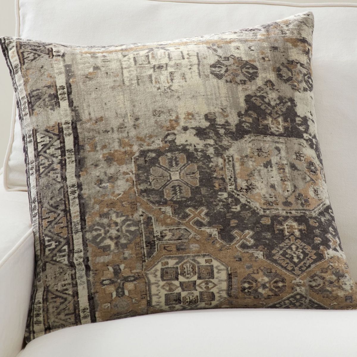 Neutral velvet throw pillow with a distressed vintage rug pattern in beige, brown, gray, and cream on a white chair.