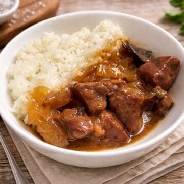 Slow cooker beef tips in gravy served over fluffy rice in a white bowl, with tender beef chunks and onions on a rustic table setting.